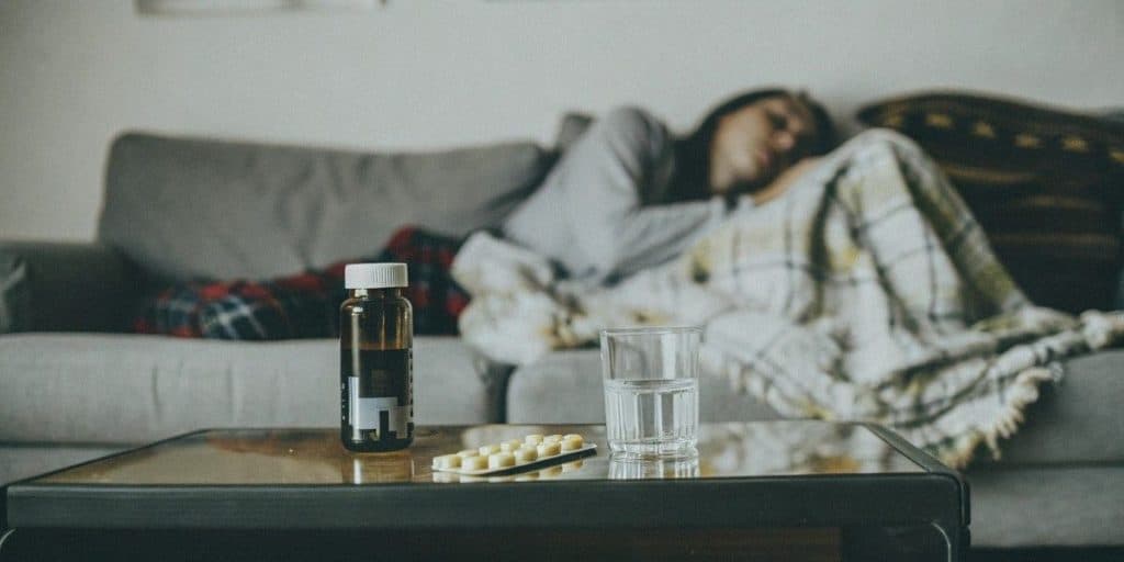 A woman feeling unwell with the flu, resting on a couch with medications placed on the coffee table next to her.